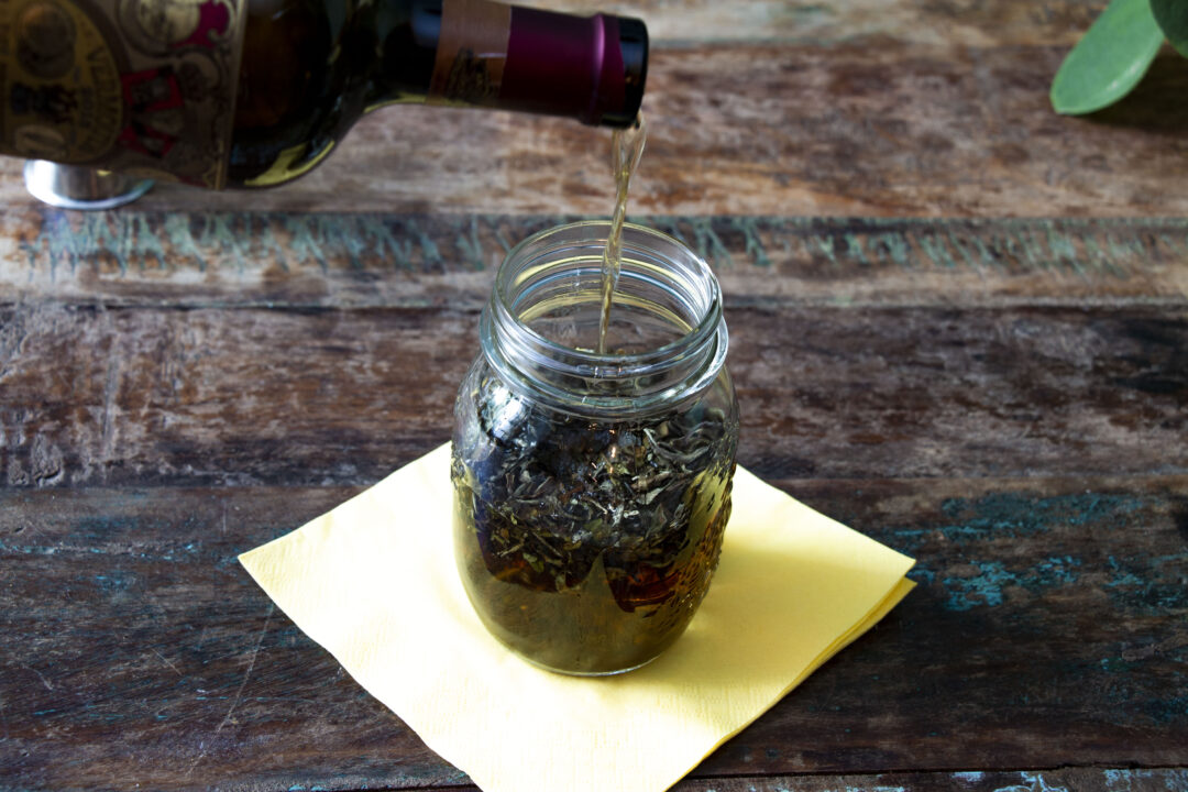A mason jar on a table with earl grey loose tea leaves inside. A bottle of sweet vermouth is pouring into the mason jar.