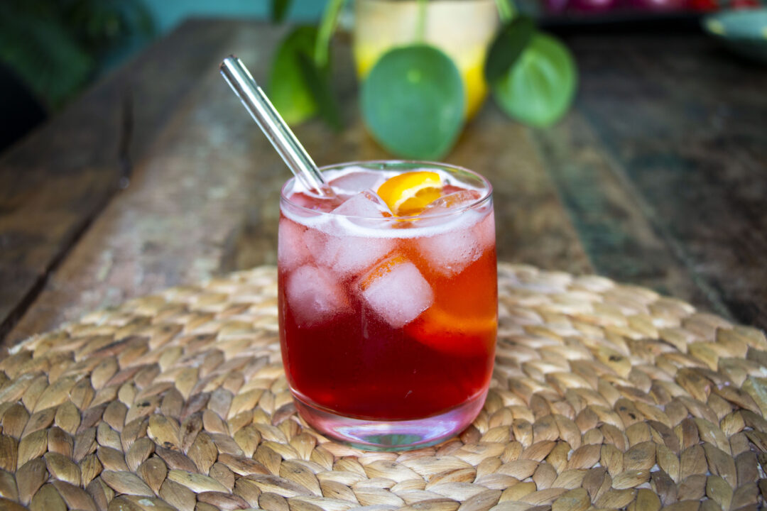 A straight on close up shot of the Caffo Spritz cocktail, which is red and bubbly with ice cubes, an orange slice and a glass straw