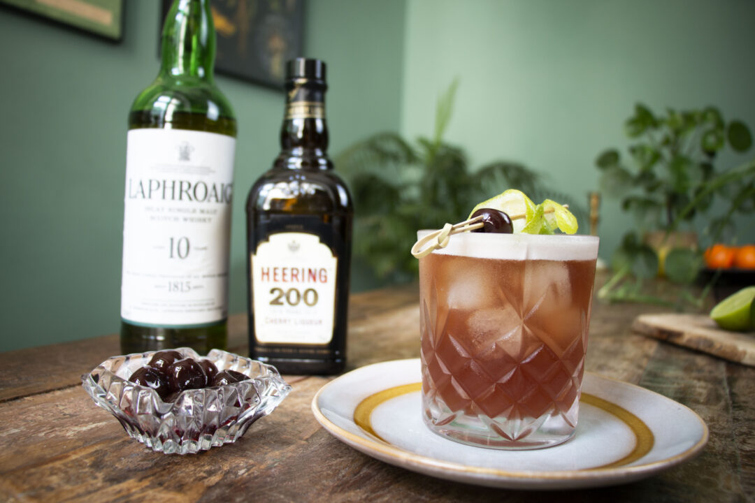 Norma Jennings cocktail on a table with bottle of Laphroaig, Cherry Heering and amarena cherries in a glass container 