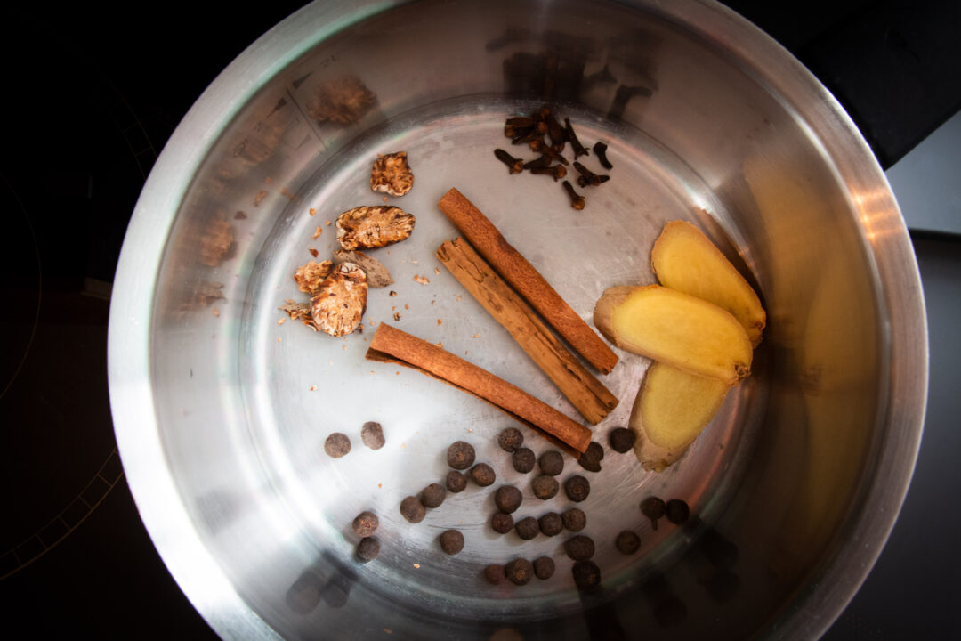 Looking down into a pot, there are five spices - fresh ginger, cinnamon sticks, cloves, a crushed whole nutmeg and allspice