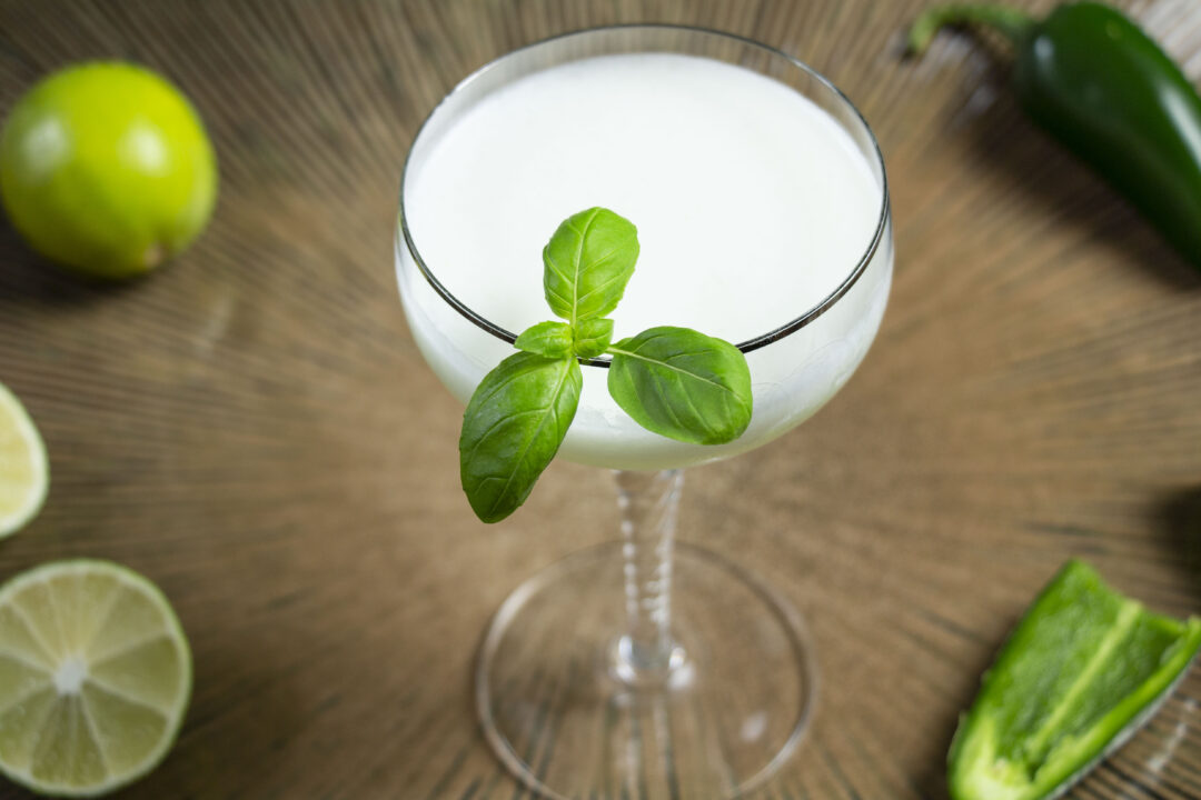 A close up overhead shot of a white cocktail with basil garnish surrounded by jalepeños and limes