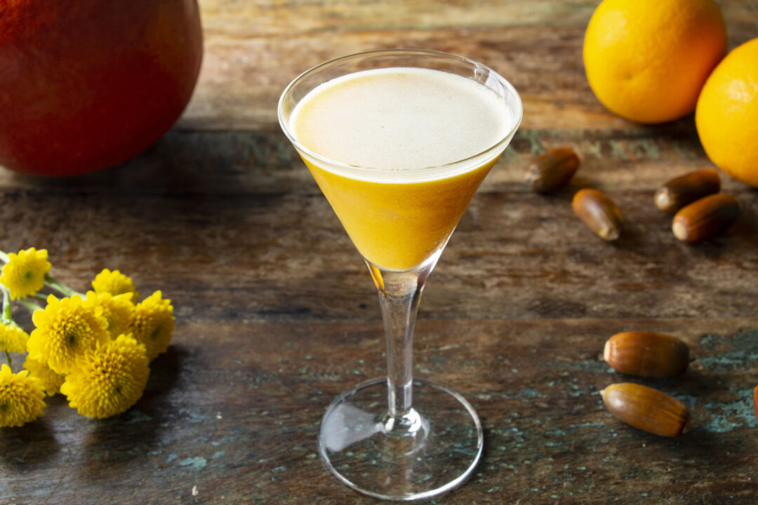 A shot of a pumpkin cocktail on a table surrounded by a pumpkin, oranges, chestnuts and yellow flowers