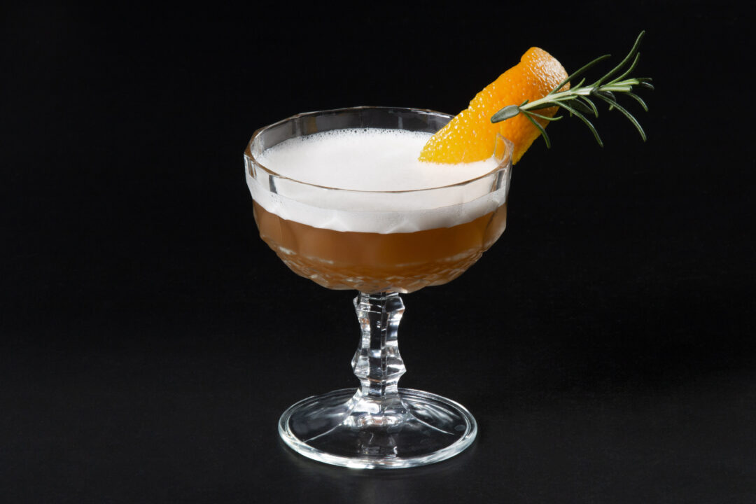 A cocktail in a stem glass with a foamy top and orange peel and rosemary garnish